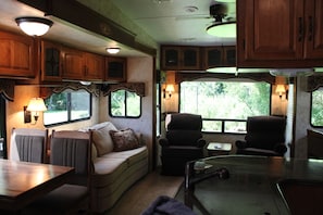 A cozy RV interior with a dining area, seating, and windows offering a view of greenery. - Cozy Mountain Camper  (Hayesville)