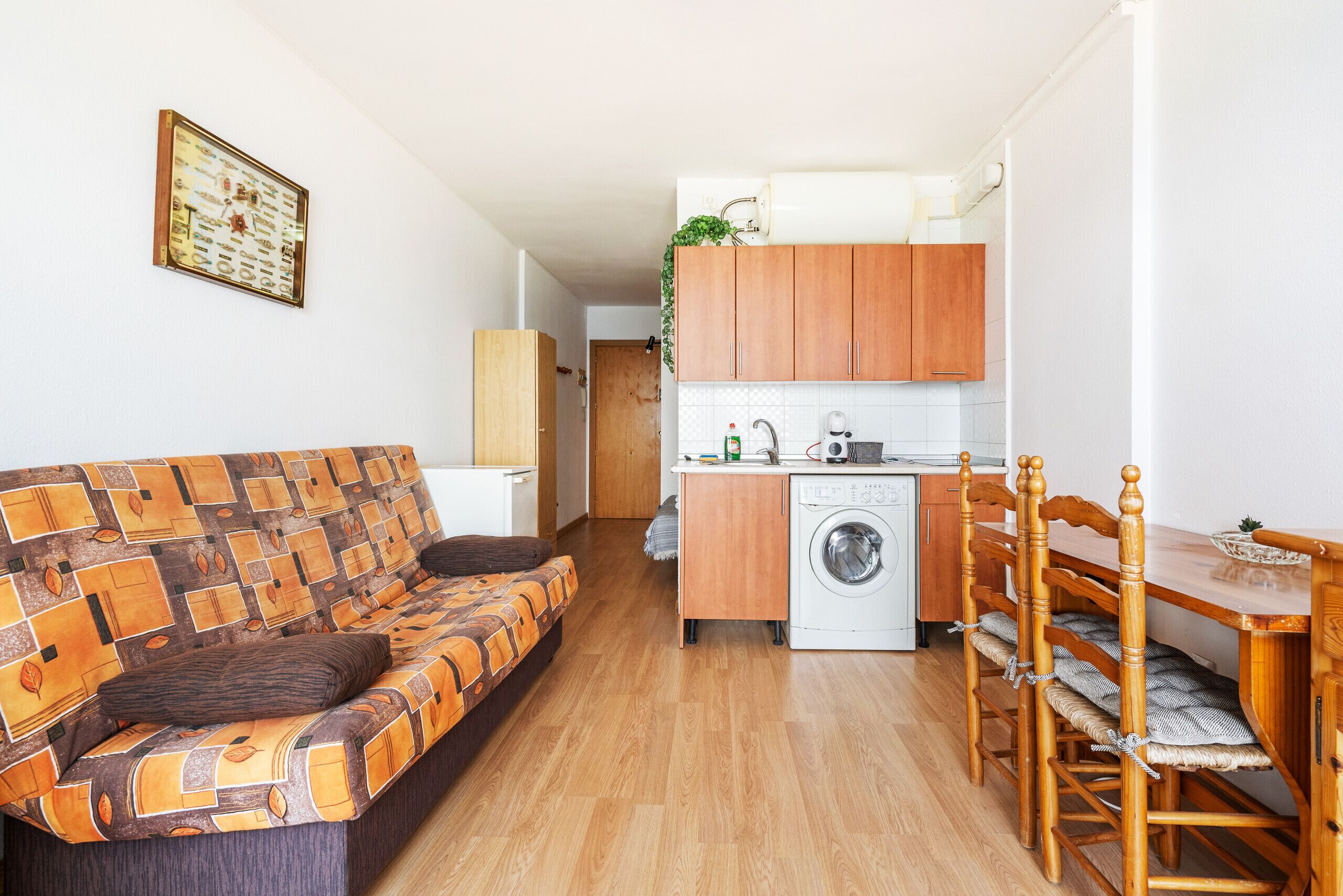 Een ruimtelijk ingerichte leefruimte met een bank, eettafel, stoelen, een wasmachine en een kitchenette.