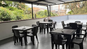 Een overdekt terras met tafels en stoelen, een betegelde vloer, en uitzicht op groen en gebouwen.