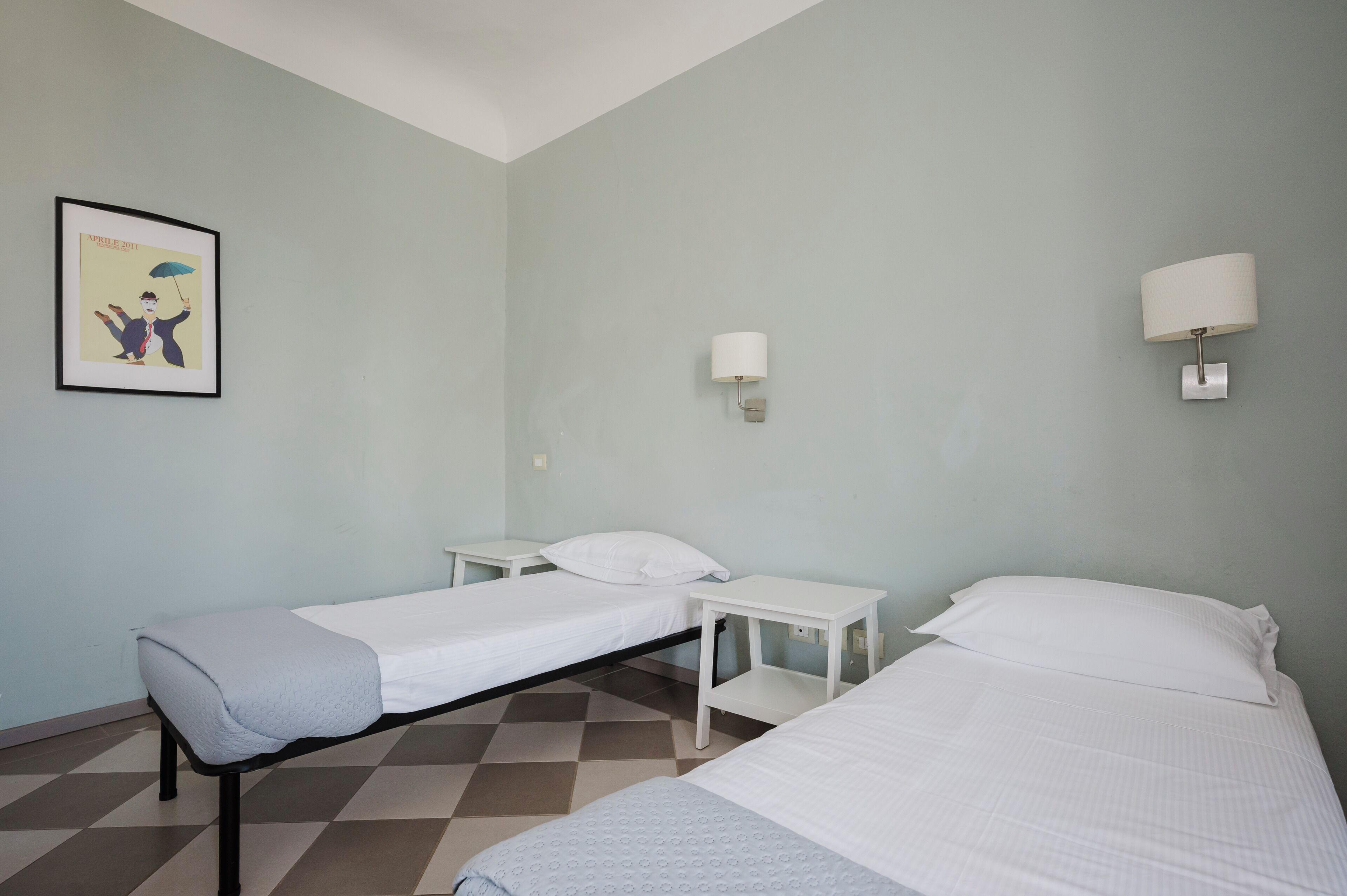 A room with two single beds, a small table, a wall-mounted lamp, and a framed picture on the wall.