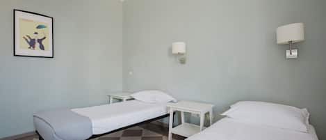 A room with two single beds, a small table, a wall-mounted lamp, and a framed picture on the wall.