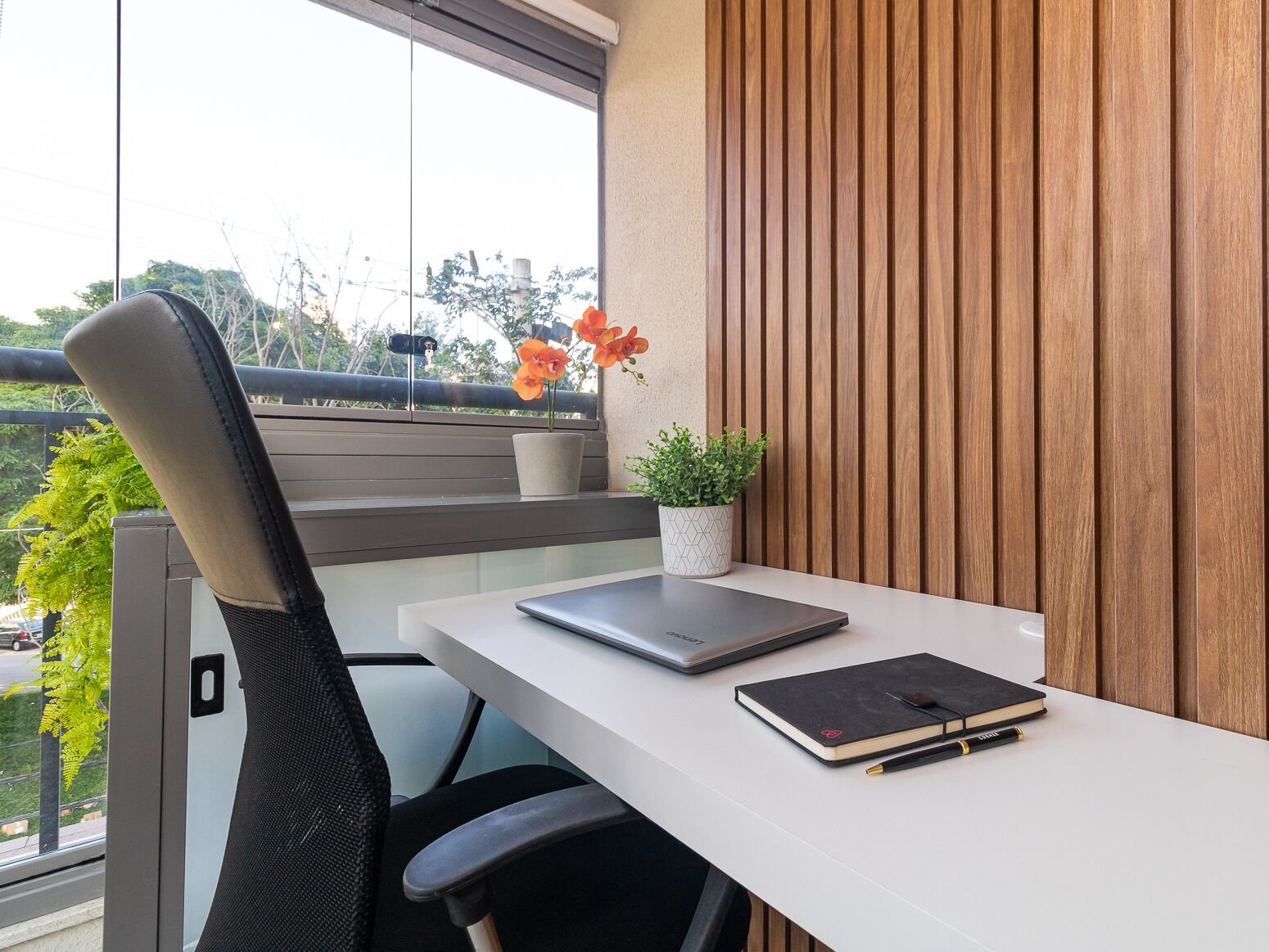 Um espaço de escritório moderno com uma mesa branca, uma cadeira de escritório preta, um laptop, um caderno e duas plantas em vasos.