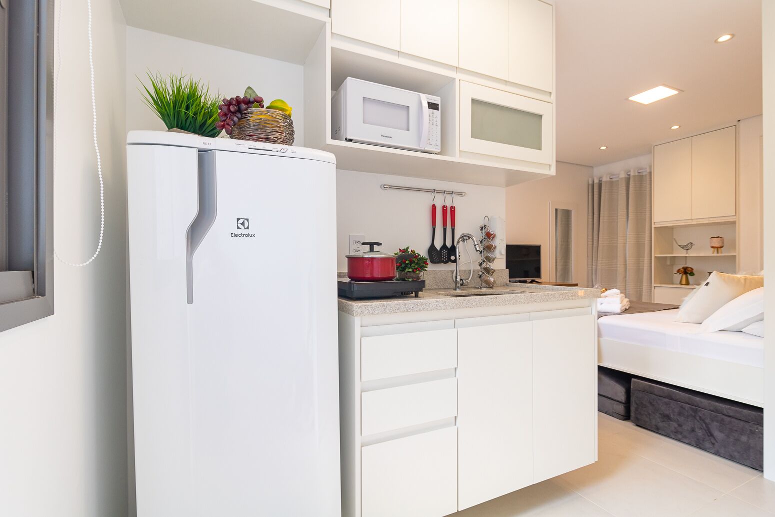 A modern kitchen with a white refrigerator, a microwave, and a sink. There is a small island with a plant and a red pot.