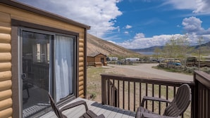 A log cabin with a deck, a covered patio, and a view of a parking area with RVs and a mountain backdrop. - Blue Mesa Recreational Ranch (Gunnison)