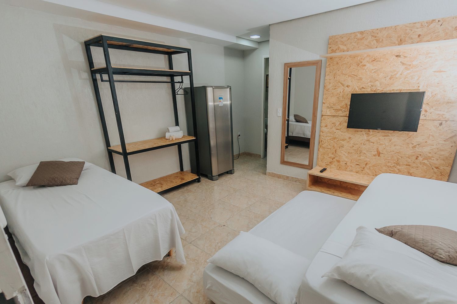 Ein Hotelzimmer mit zwei Einzelbetten, einem Kleiderschrank, einem Kühlschrank, einem Spiegel und einem an einer Holzwand befestigten Fernseher.