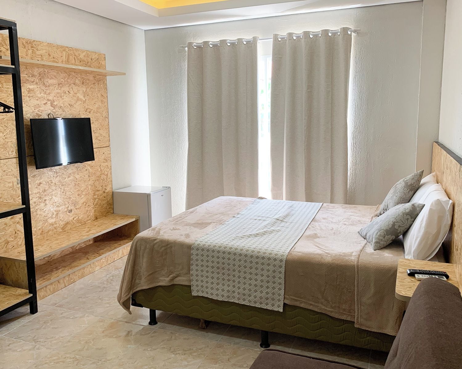 A bedroom with a bed, a TV mounted on a wooden wall, a bedside table with a remote, and a window with curtains.