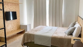 Quarto com cama, televisão presa na parede de madeira, mesa de cabeceira com controle remoto e janela com cortinas.