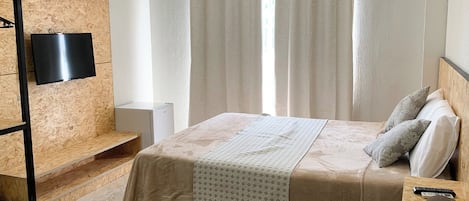 A bedroom with a bed, a TV mounted on a wooden wall, a bedside table with a remote, and a window with curtains.