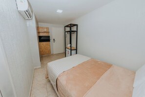 A narrow room with a bed, a small kitchenette, and a door leading to another room.