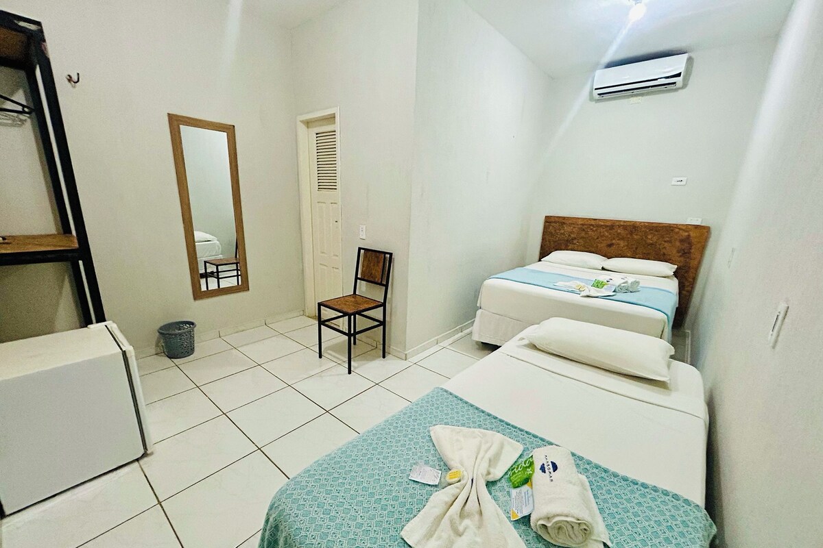 A hotel room with a bed, a chair, a small table, a mirror, and an air conditioning unit.