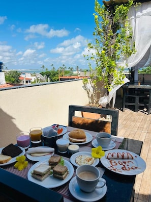 Una tavola apparecchiata con diversi cibi per la colazione, tra cui panini, pasticcini e bevande, con vista su una zona costiera con palme e cielo sereno.