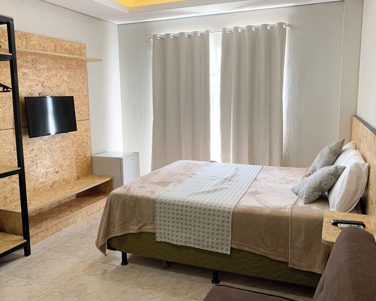 Ein Schlafzimmer mit einem Bett, einem an einer Holzwand montierten Fernseher, einem Nachttisch mit Fernbedienung und einem Fenster mit Vorhängen.