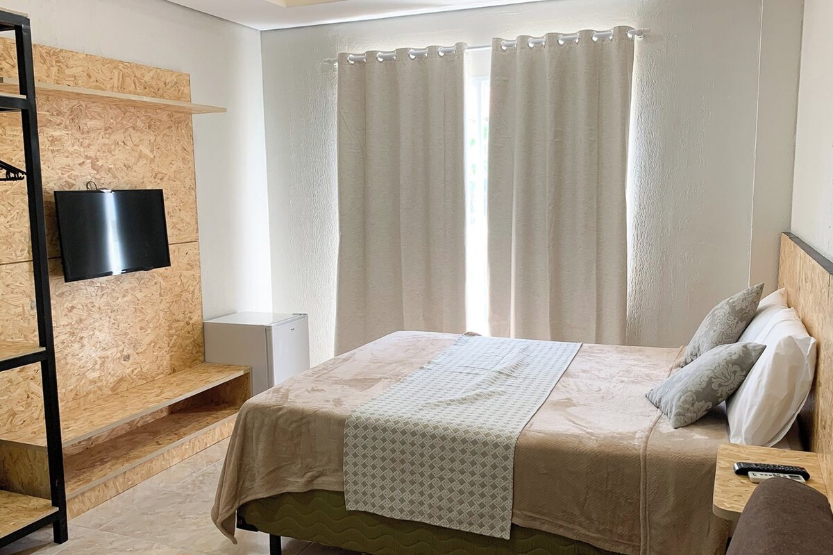A bedroom with a bed, a TV mounted on a wooden wall, a bedside table with a remote, and a window with curtains.