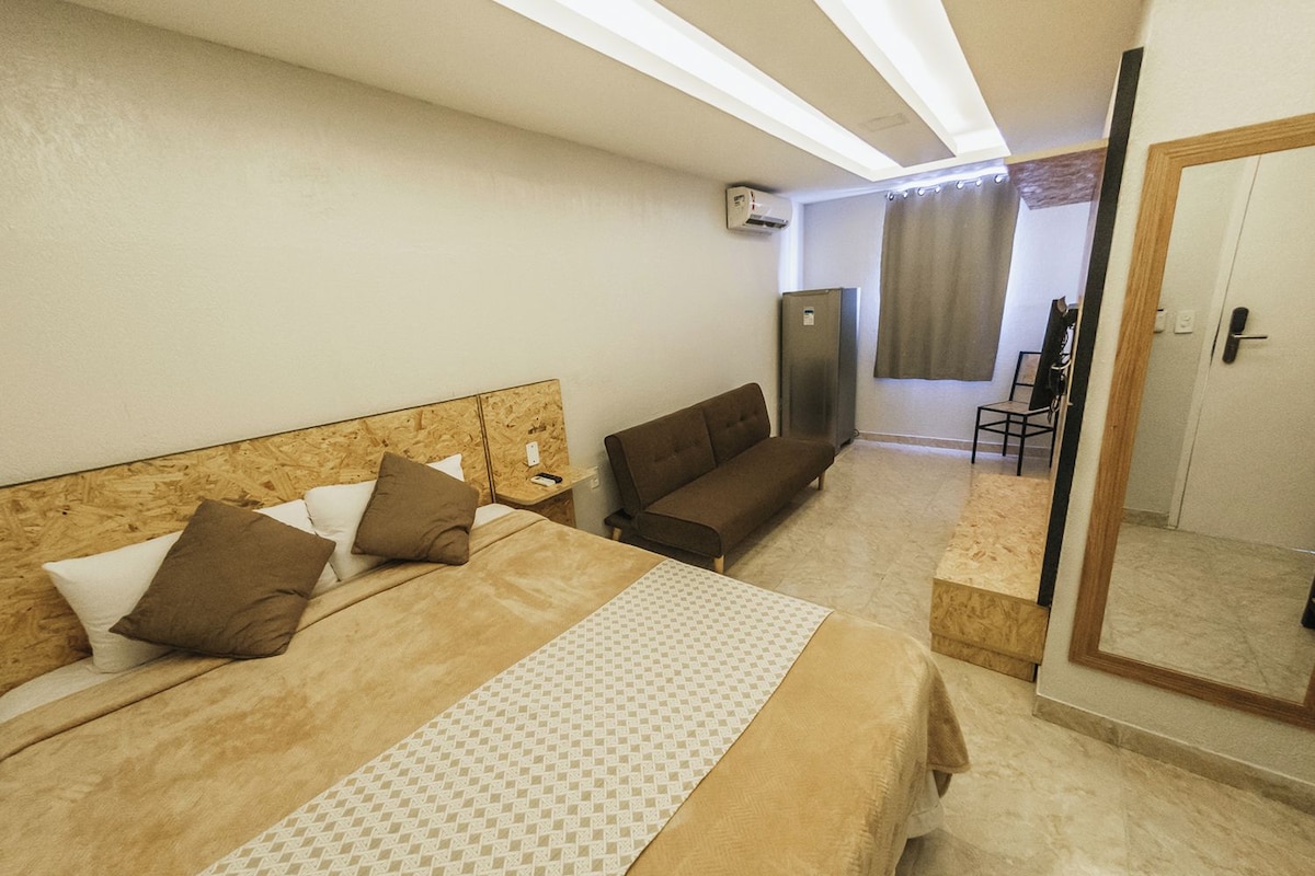 A hotel room with a bed, a sofa, a mirror, and a refrigerator.