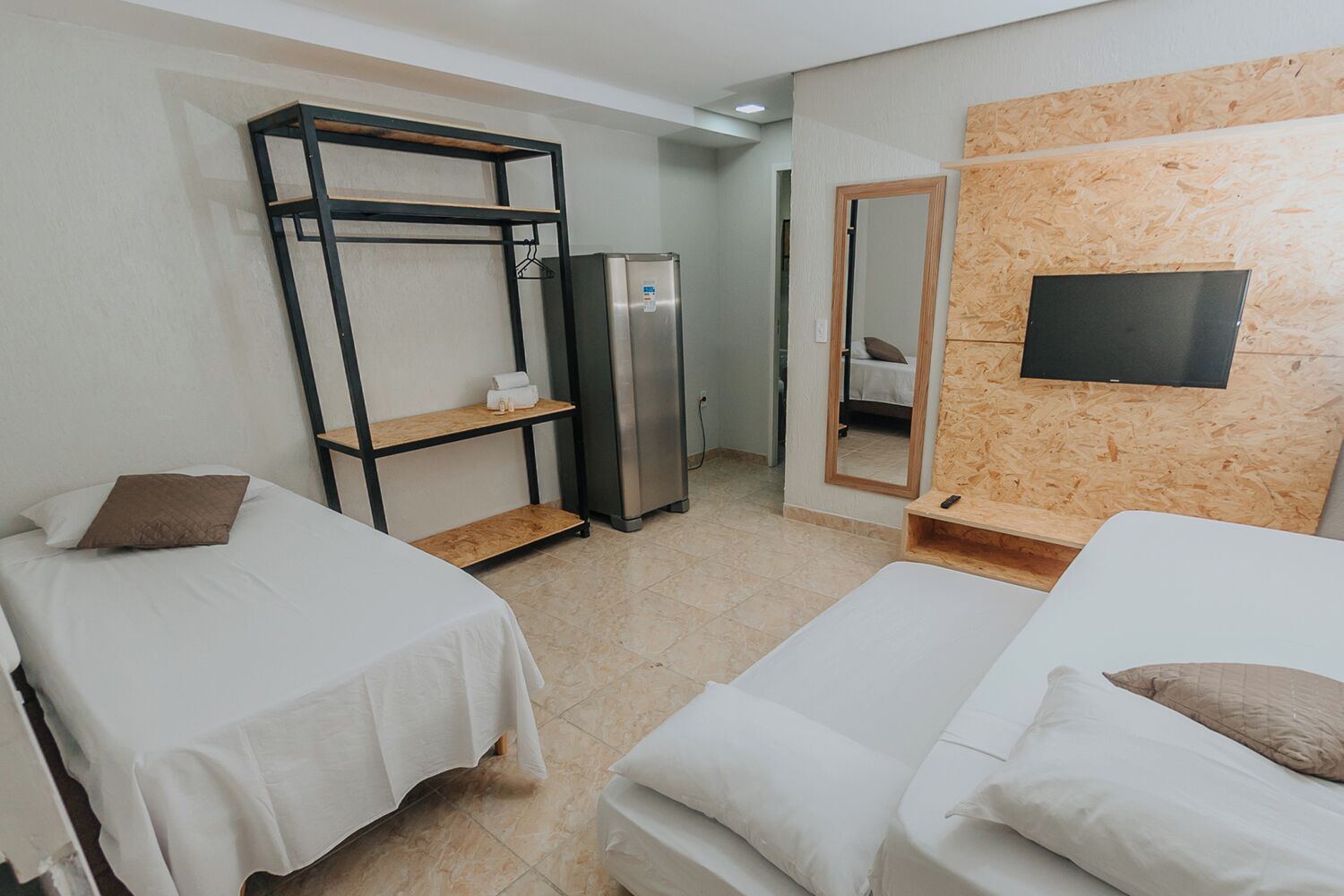 Una habitació d'hotel amb dos llits individuals, un armari, un refrigerador, un mirall i una televisió penjada a una paret de fusta.