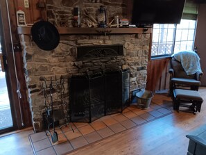 Una chimenea de piedra con un televisor encima, un sillón de cuero con una manta, y piso de madera.