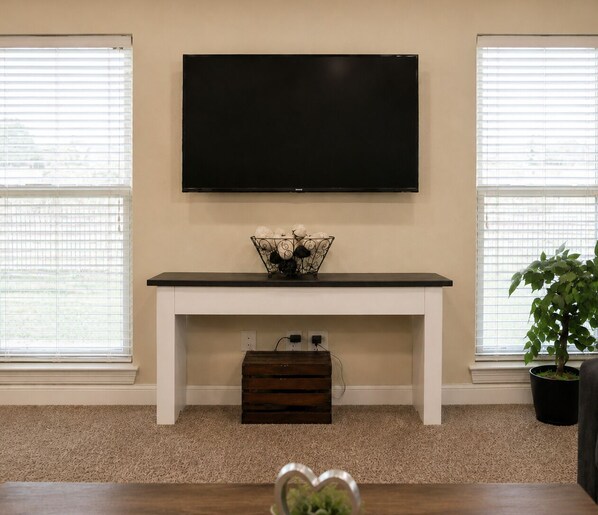 A living room with a TV mounted on the wall, a white console table, a decorative basket, a potted plant, and a wooden chest of drawers. - Work Hard, Rest Easy · 3BR 2BA · W/D · Gulf Coast (Moss Point)