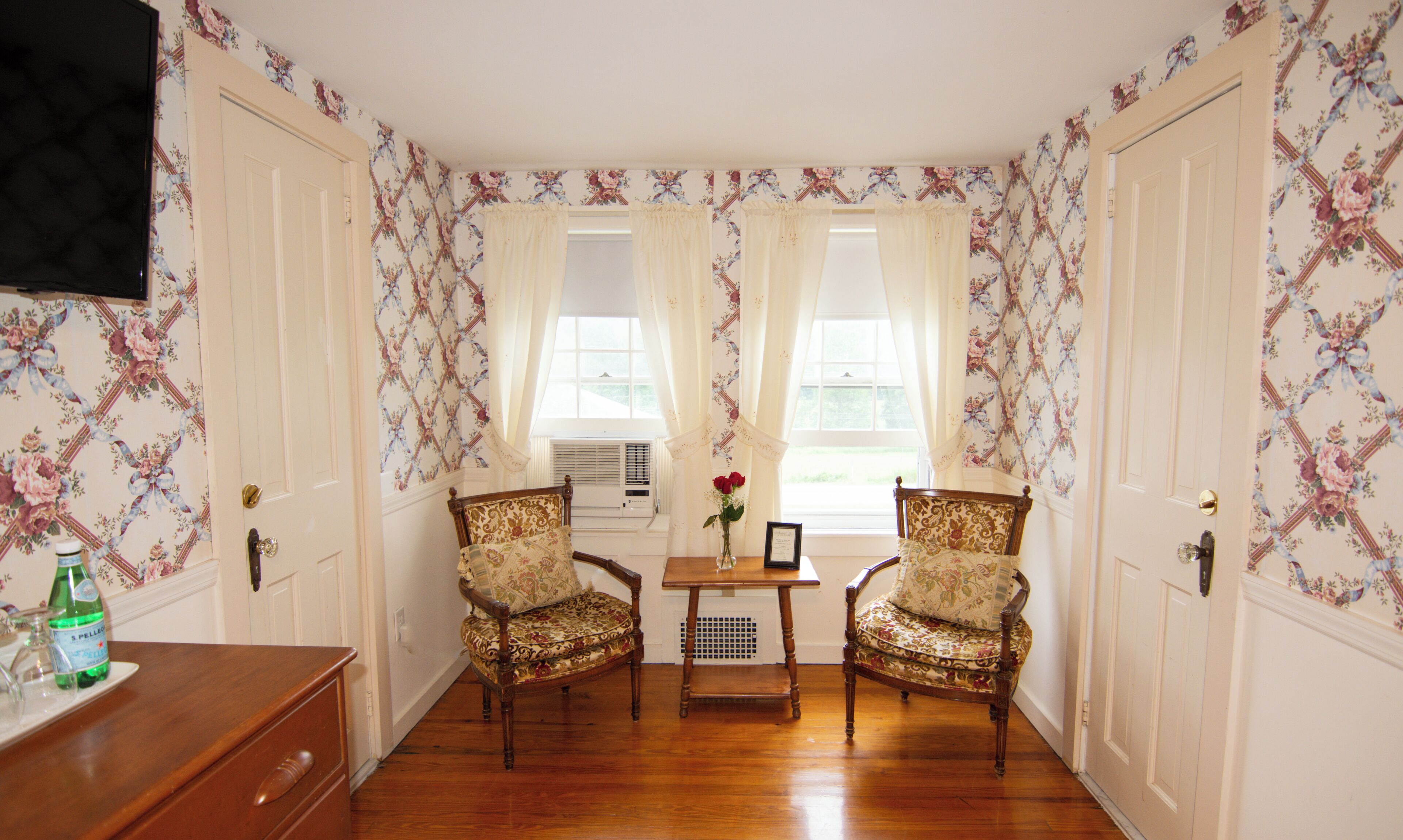 A room with floral wallpaper, two upholstered chairs, a small table with a vase and a picture frame, and a wooden dresser.