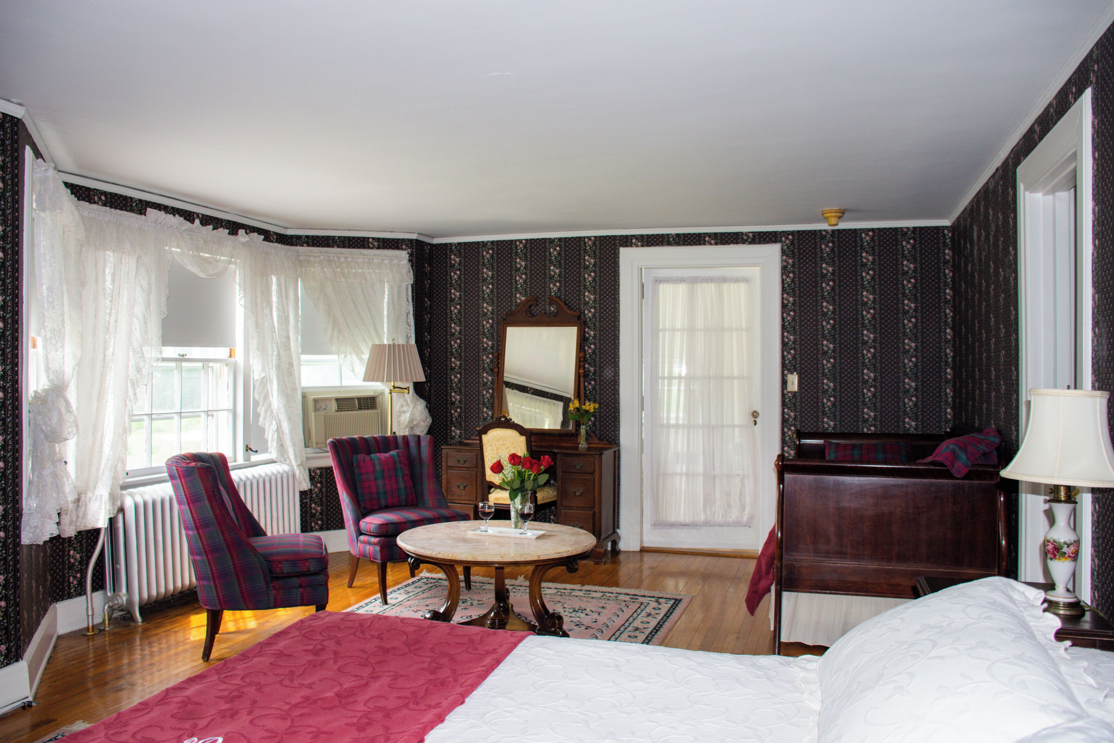 A bedroom with a large bed, a round table with a vase of flowers, a chair, a dresser, and a mirror.
