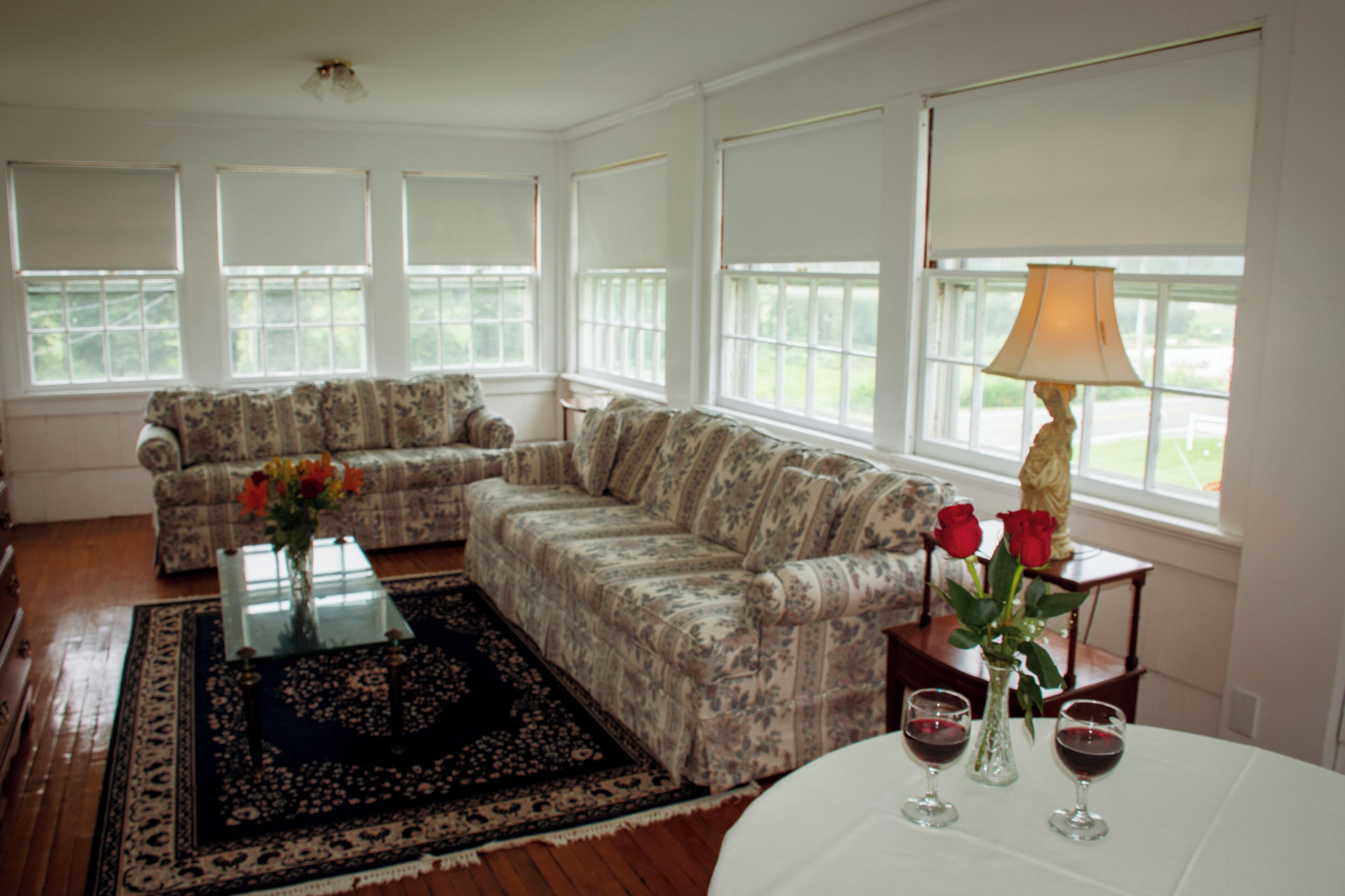 Una sala de estar con un sofá floral, una mesa de centro de vidrio y un jarrón con flores. Hay dos copas de vino sobre una mesa.