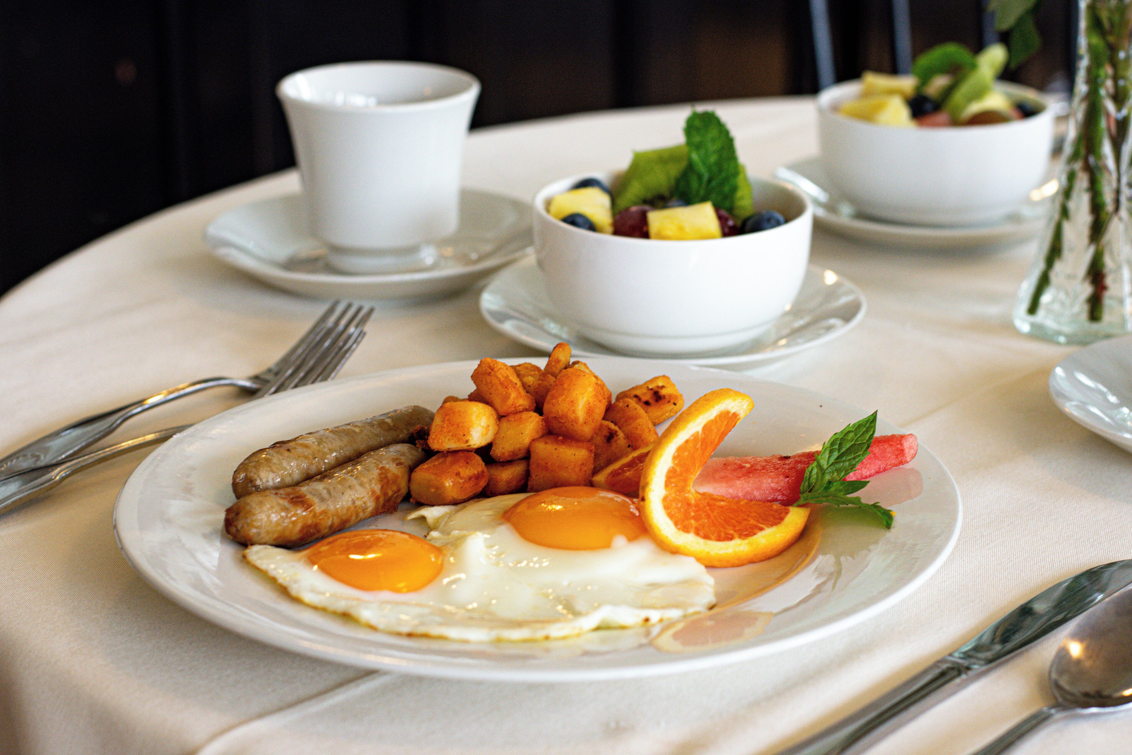 A breakfast setting with plates of eggs, sausages, and potatoes, accompanied by bowls of fruit and a cup.