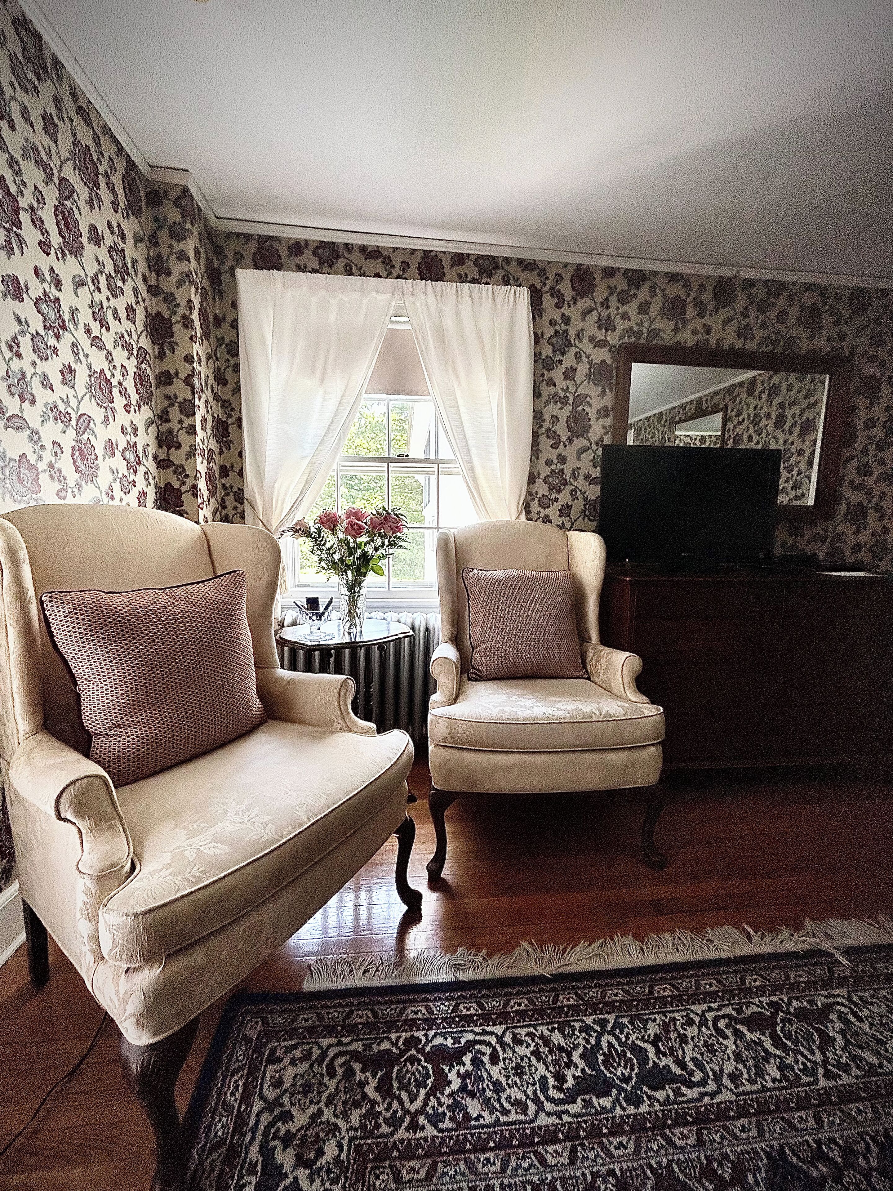 A living room with two armchairs, a small table with a vase of flowers, a television, and patterned wallpaper.