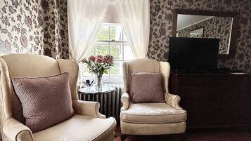 A living room with two armchairs, a small table with a vase of flowers, a television, and patterned wallpaper.