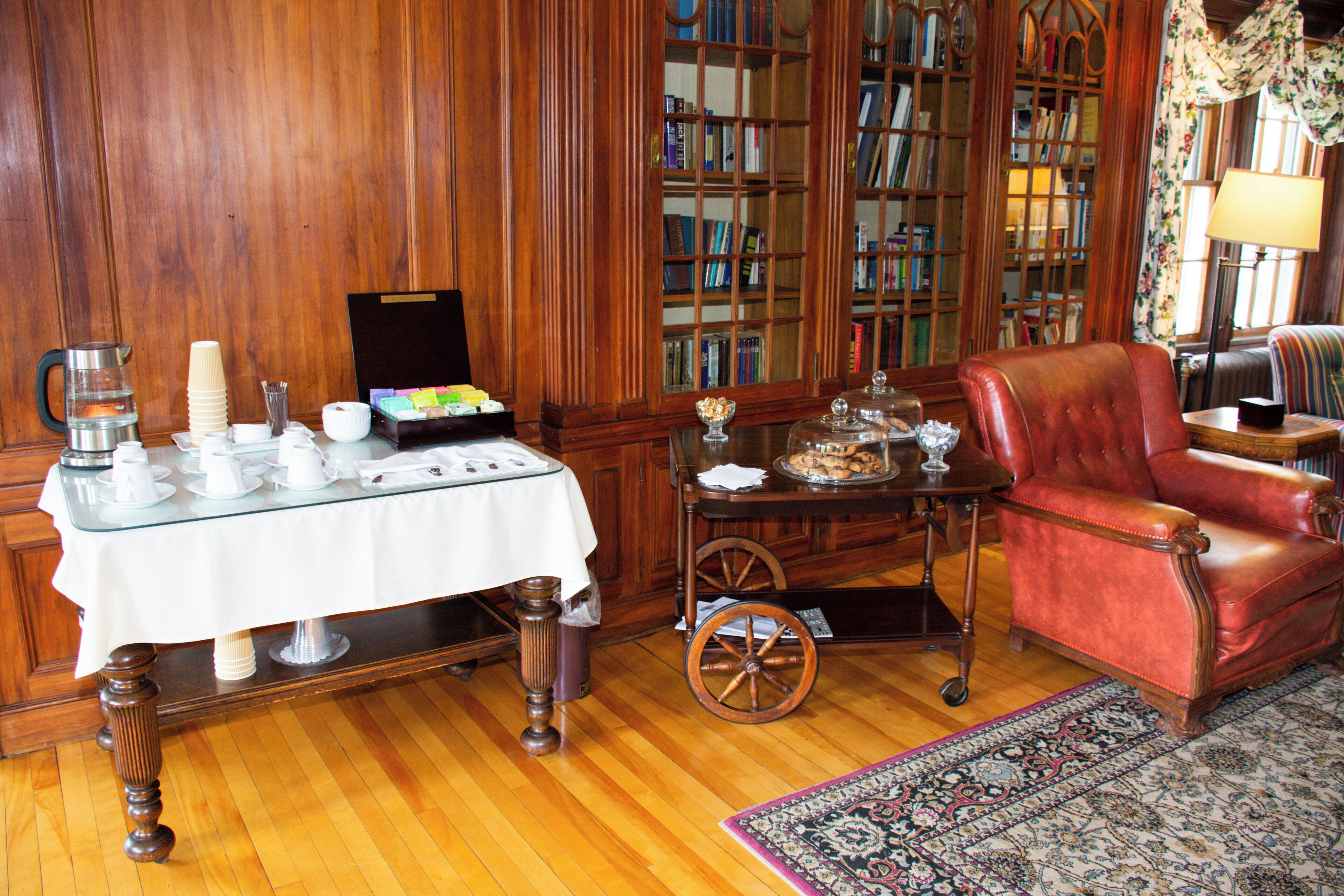 Una habitación con piso de madera, un sillón de cuero rojo, una mesa con mantel blanco y una estantería llena de libros.