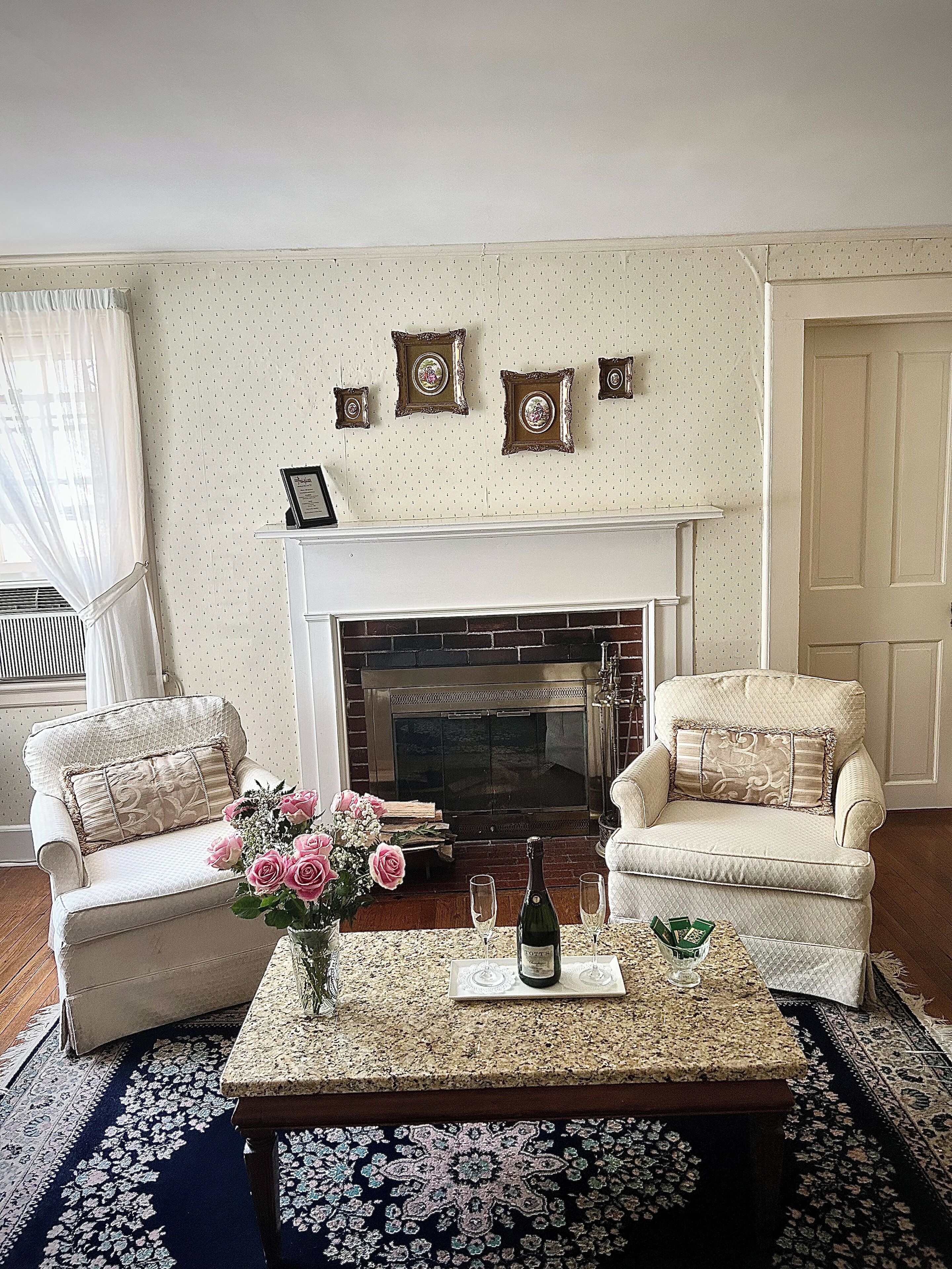 A living room with a fireplace, two armchairs, a coffee table with a vase of flowers, and a framed picture collection on the wall.