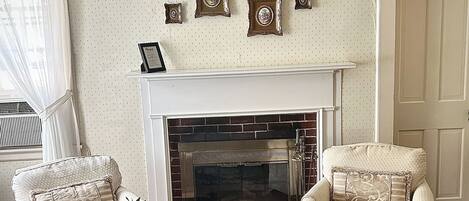 A living room with a fireplace, two armchairs, a coffee table with a vase of flowers, and a framed picture collection on the wall.