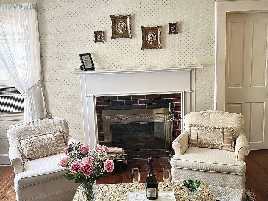 Ett vardagsrum med en öppen spis, två fåtöljer, ett soffbord med en vas med blommor och en vägg dekorerad med en samling inramade bilder.