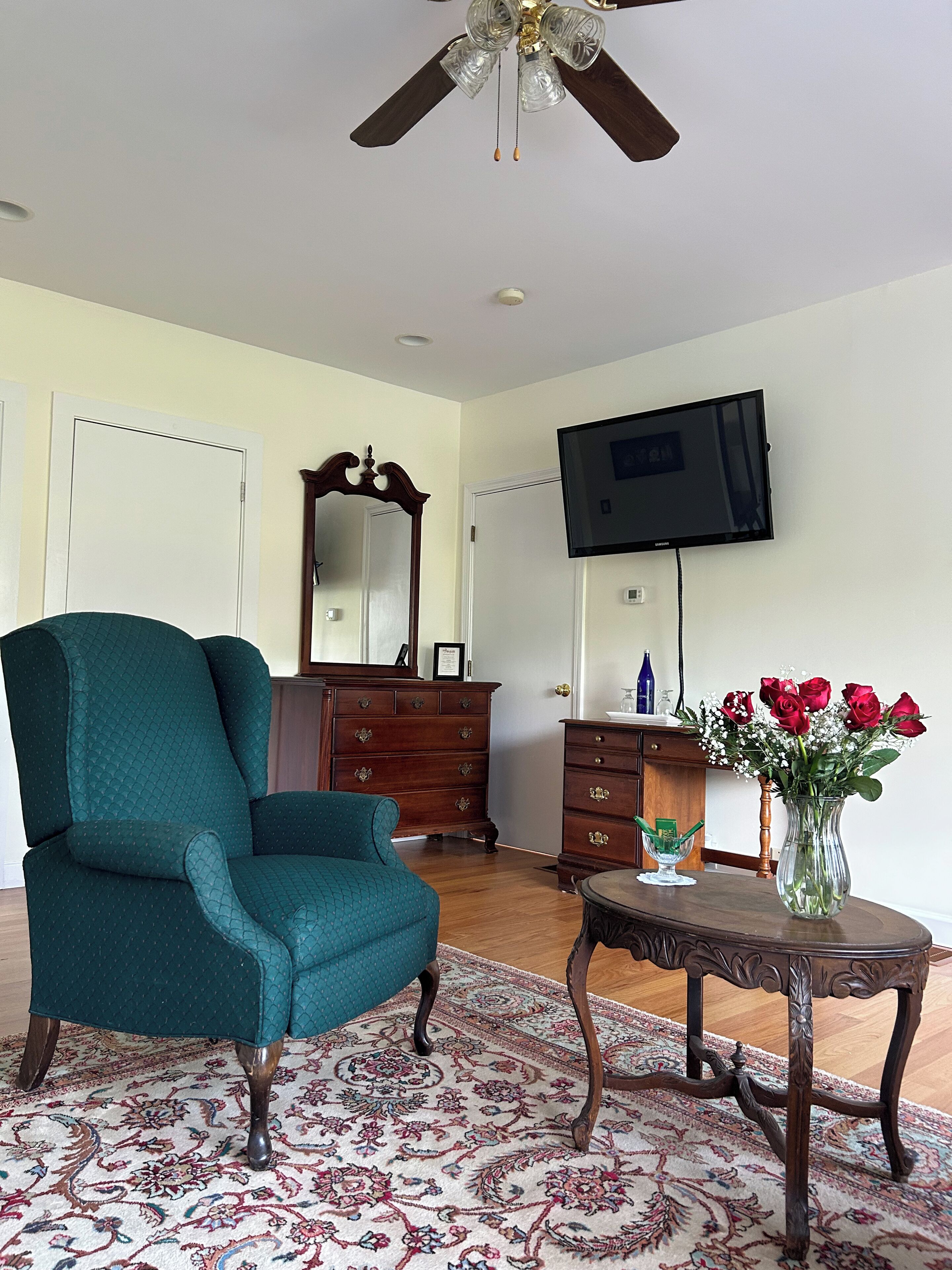 Uma sala de estar com uma poltrona azul-turquesa, um aparador de madeira, uma televisão montada na parede e um vaso com rosas vermelhas sobre uma mesa lateral.