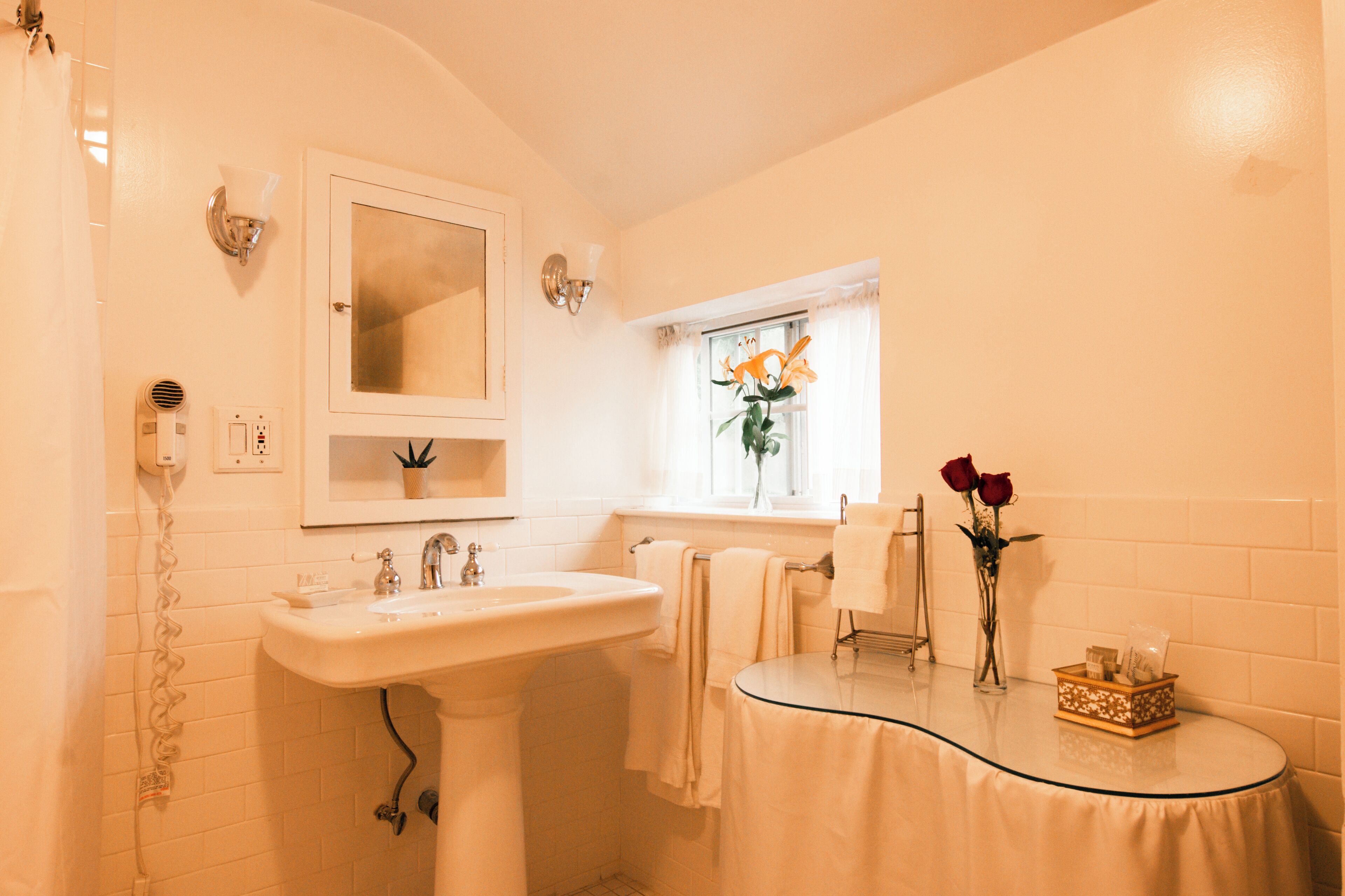 A bathroom with a sink, mirror, window, and a small table with a vase of flowers.