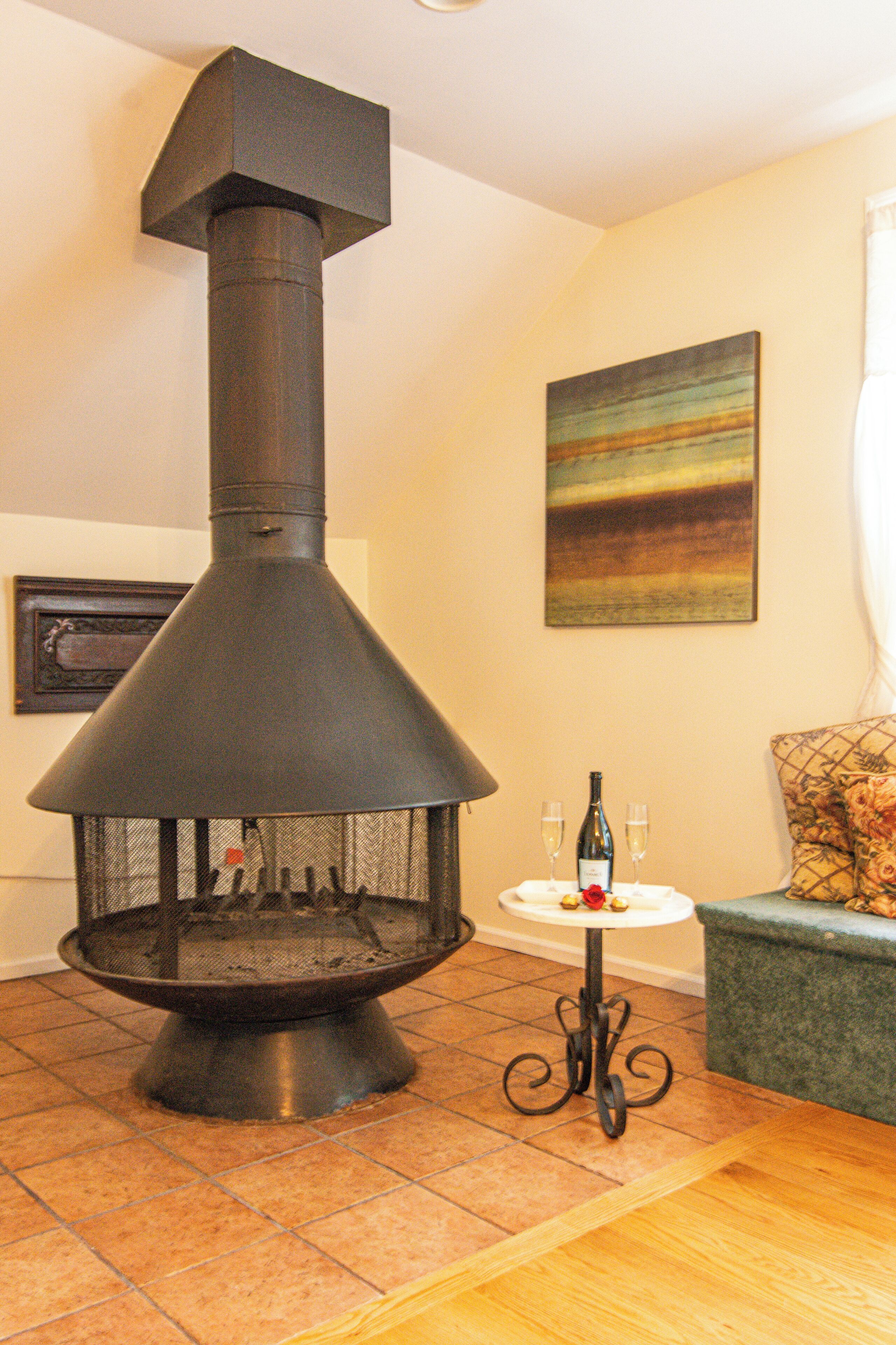 Un estufa de leña con chimenea, una mesita con una botella y vasos, y un sofá con un cojín estampado.