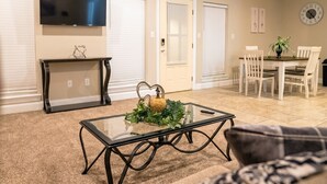 A living room with a glass-top coffee table, a TV mounted on the wall, a fireplace, a dining area with chairs, and a clock on the wall. - Contractor's Choice · 3BR · Near Ingalls/Chevron (Moss Point)