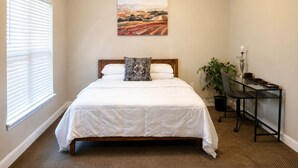 Un dormitorio con cama, escritorio, silla, planta en maceta y un cuadro en la pared.