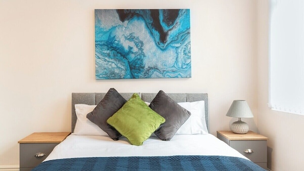 Ein Schlafzimmer mit einem Bett, Kissen, einem Nachttisch, einer Lampe und einem Wandbild.