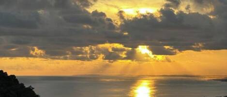 A coastal town at sunset with the sun setting over the water, surrounded by clouds and buildings.