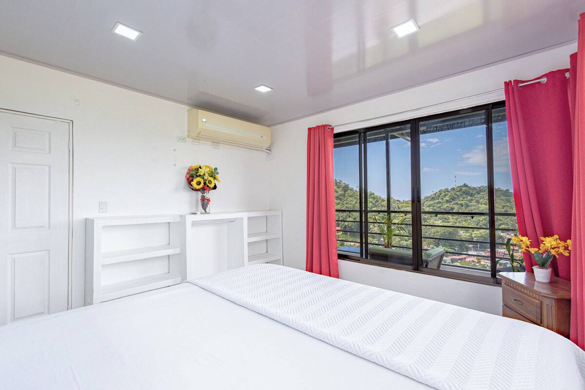 A bedroom with a large bed, a window with curtains, a desk with a vase of flowers, and a view of greenery outside.