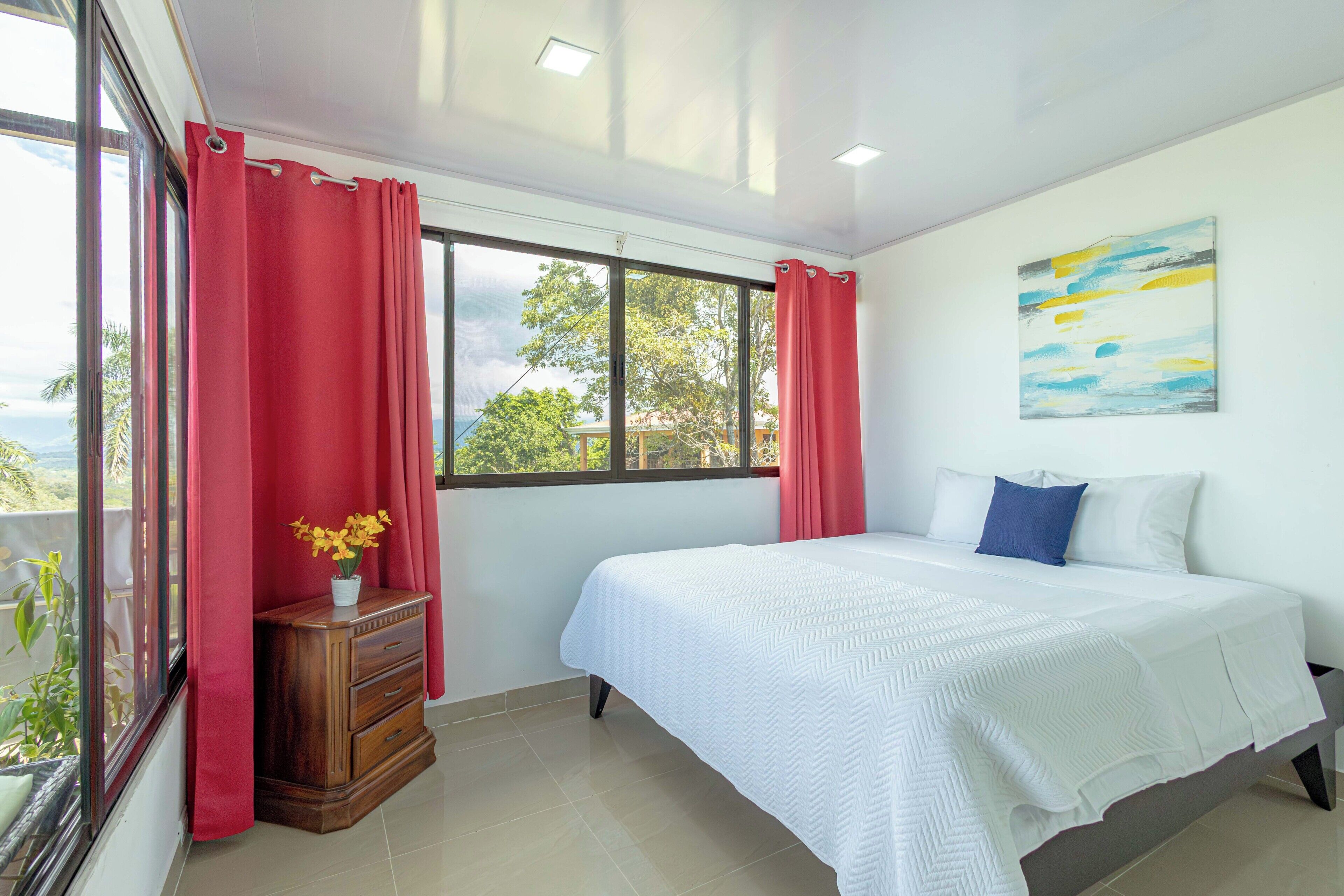 Un dormitorio con una cama, una ventana con cortinas, un cuadro en la pared y una cómoda de madera con un jarrón de flores.