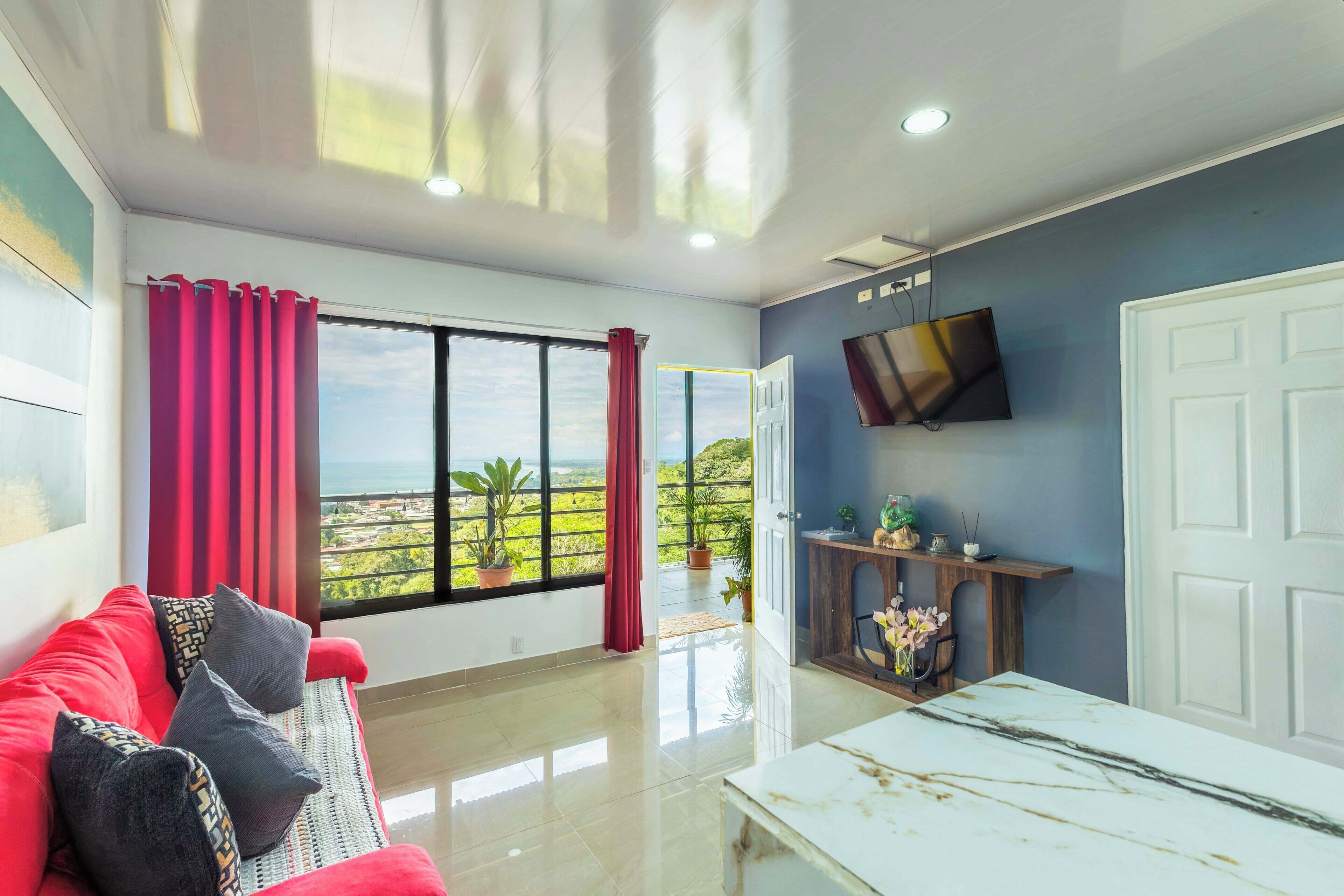 A modern living room with a red sofa, a white coffee table, a flat-screen TV, and a large window with curtains.
