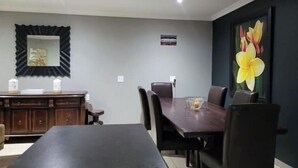 Een eetgedeelte met een lange tafel, stoelen, een houten kast en een groot bloemschilderij aan de muur.