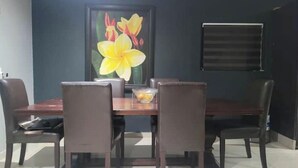 Een eetgedeelte met een donkerhouten tafel en vier stoelen, een wanddecoratie met bloemen in een lijst en een raam met zonwering.