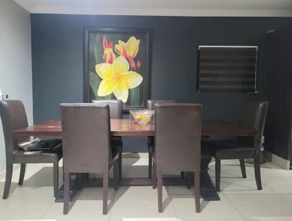A dining area with a dark wooden table and four chairs, a framed floral artwork on the wall, and a window with blinds. - Golf n Big5 rural experience (uMhlanga)