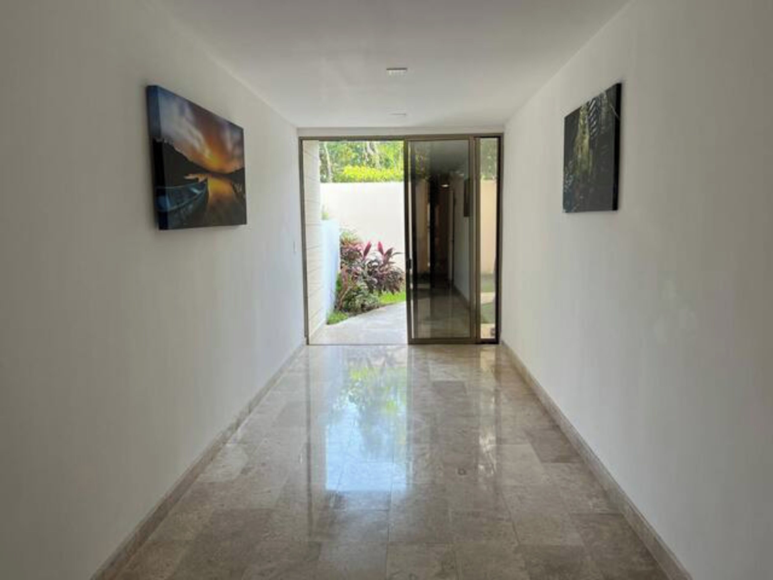 A hallway with a glass door leading to an outdoor area, flanked by two artworks on the walls.