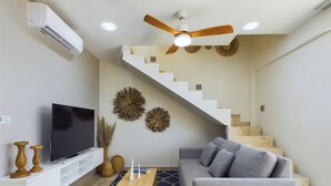 Moderni olohuone, jossa on harmaa sohva, litteänäyttöinen televisio, puinen sohvapöytä ja portaikko, jonka yläpuolella on puinen kattotuuletin.