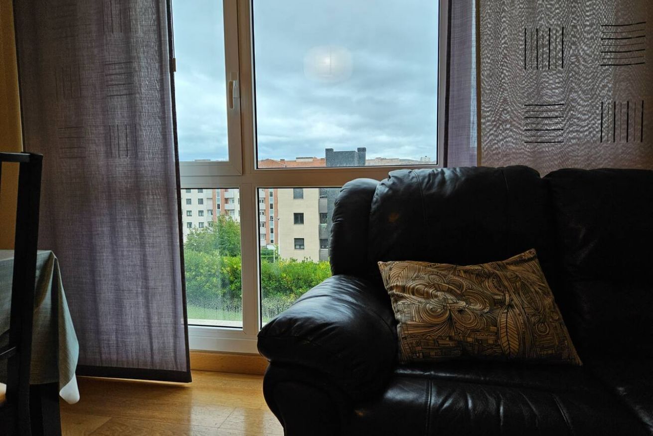 A room with a black leather sofa, a patterned pillow, a window with a view of buildings and greenery, and a curtain partially drawn.