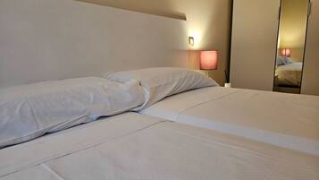 Una cama bien hecha con sábanas blancas, una lámpara de noche y un espejo que refleja la habitación.