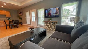 A living room with a dark coffee table, a gray sofa, and a TV displaying a movie. There is a dining area with wooden cabinets and a chair. - Unwind After Work · 3BR 2BA · King Beds · W/D (Moss Point)