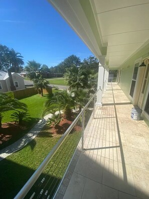 A balcony with a view of a green lawn and palm trees, a tiled floor, and a metal railing. - Unwind After Work · 3BR 2BA · King Beds · W/D (Moss Point)